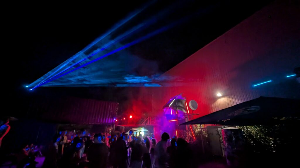 Outdoor night dancefloor under a canopy, with blue laser beams cutting through red-lit haze