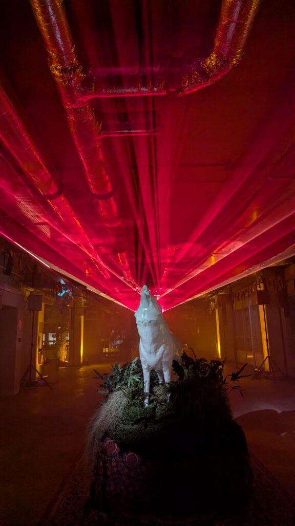 White wolf sculpture on a mossy plinth, lit from above, with red laser beams across the ceiling