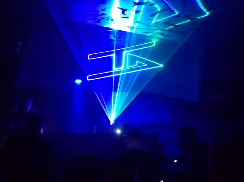 Angular white logo drawn in the air by lasers, above converging cyan and blue beams over a crowd